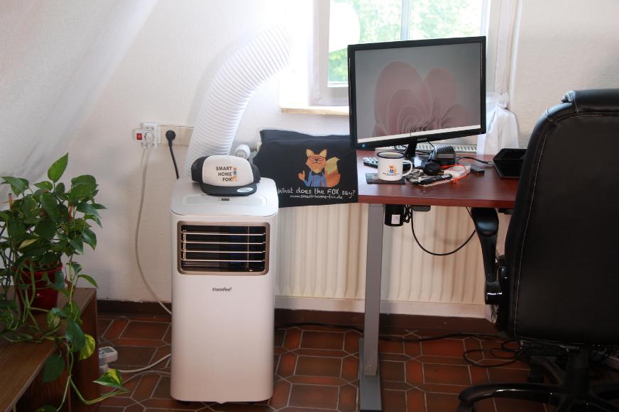 Eine mobile Klimaanlage mit Abluftschlauch steht im Büro und der Abluftschlauch hängt durch eine Fensterabdichtung nach draußen.